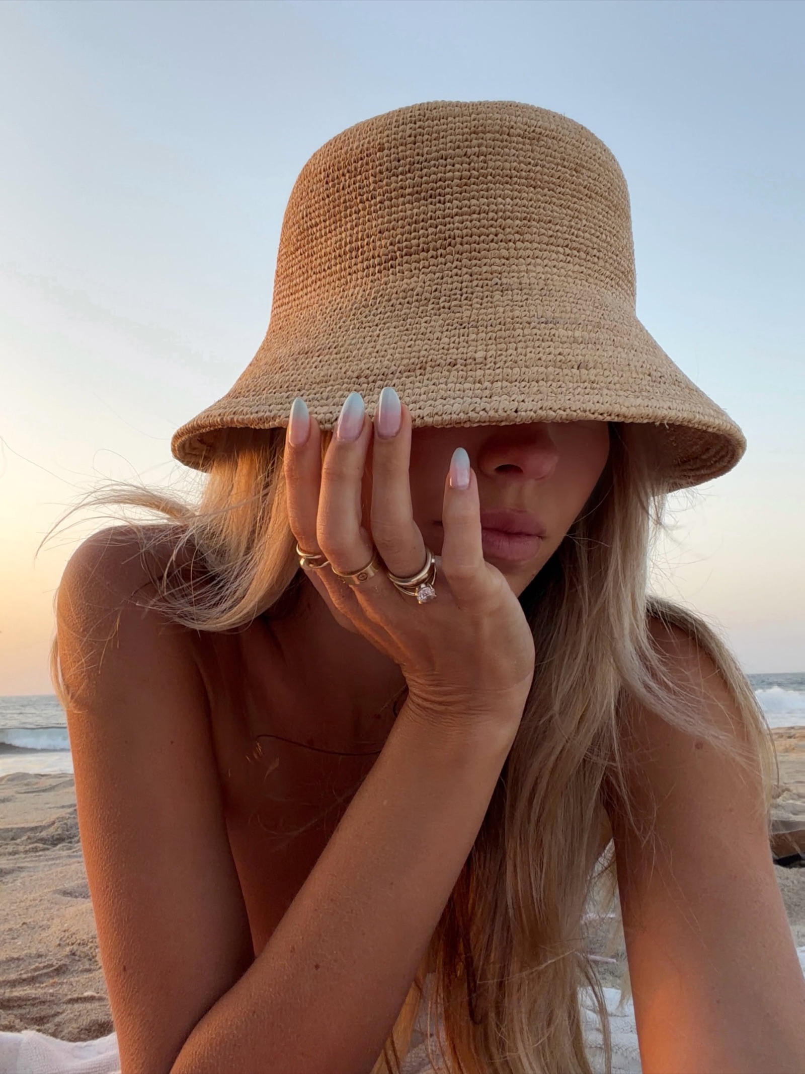 Woman in a straw bucket hat hiding her face on the beach at sunset